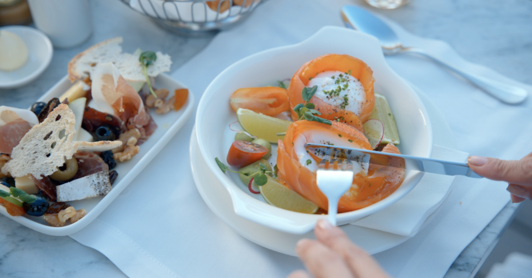 Captura de pantalla 2026-10-26 a la(s) 5.47.29 p.m. Person cuts into smoked salmon and burrata salad with lime and herbs, beside a platter of cheese, olives, cured meats, and bread.