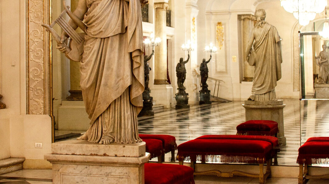 Musa-foyer-agustinas_optimizada Classical marble statue in an ornate mirrored hall with crystal chandeliers, red velvet benches, and a black-and-white checkerboard floor.