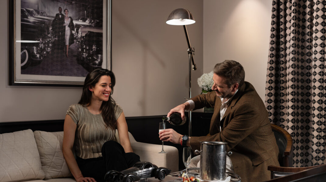 NZ6_8630-Edit A man pours champagne for a smiling woman seated on a sofa in an elegant lounge.