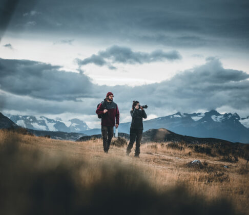 _SEB7224 Two hikers cross a golden alpine meadow beneath dramatic storm clouds, with snow-capped mountains in the distance.