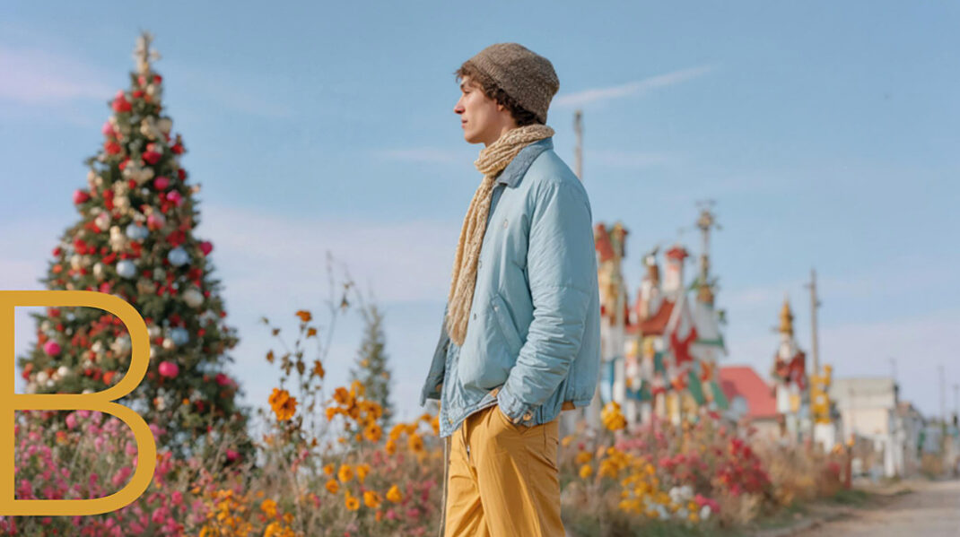 Teatro_M_2_optimizada Man wearing a beanie, scarf, and blue jacket stands on quad roller skates beside a decorated Christmas tree and colorful flowers.