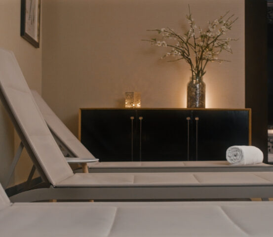 expwellstgo_web_HQ Warmly lit spa lounge with cushioned chaise lounges, a rolled towel, flowers, and a tiled soaking tub.