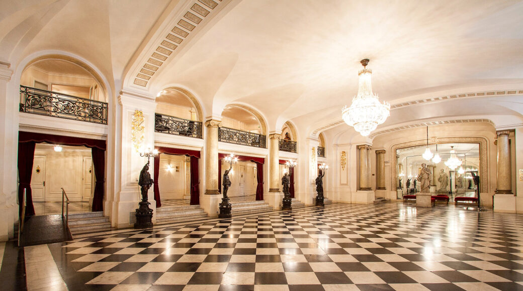 foyer-agustinas_optimizada Ornate palace hall with a black-and-white checkerboard floor, crystal chandelier, marble columns, and bronze statues.