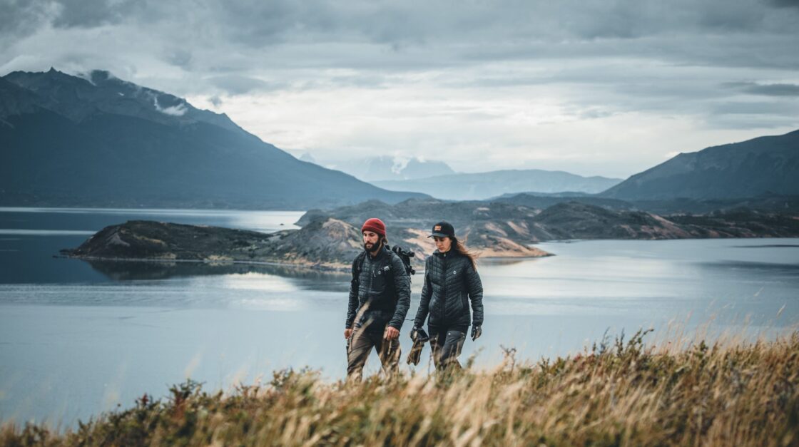 hon1 Two hikers in outdoor jackets walk above a lake, with rugged mountains beneath a cloudy sky.