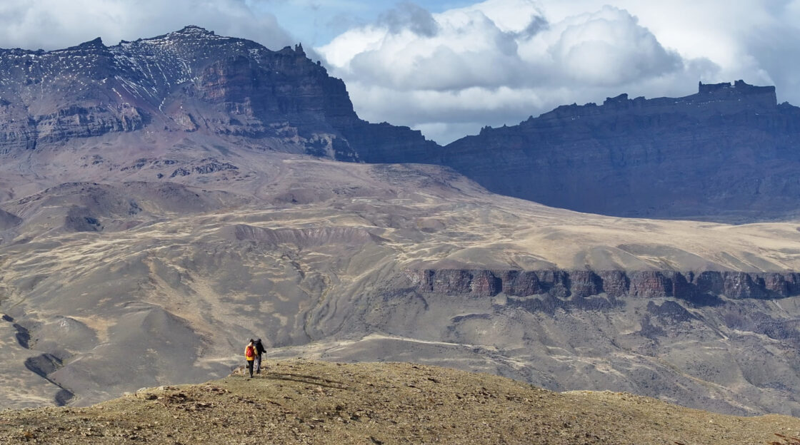 Screenshot Two hikers traverse a barren alpine ridge beneath towering, snow-dusted mountains and low clouds.