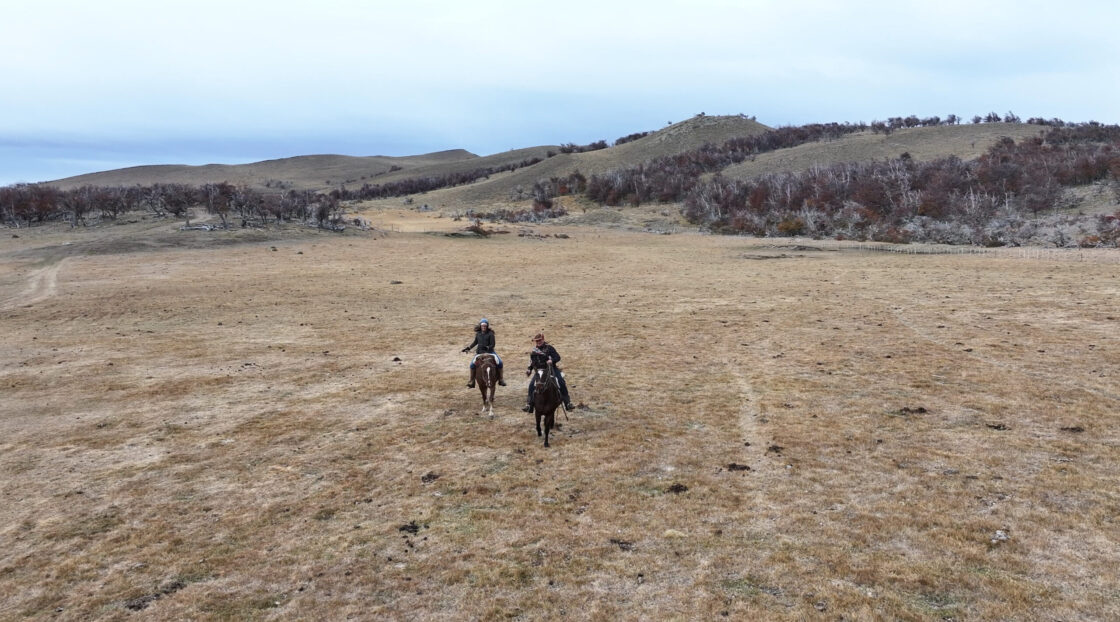 Screenshot Two riders on horseback cross a dry grassland beneath rolling hills and sparse trees.