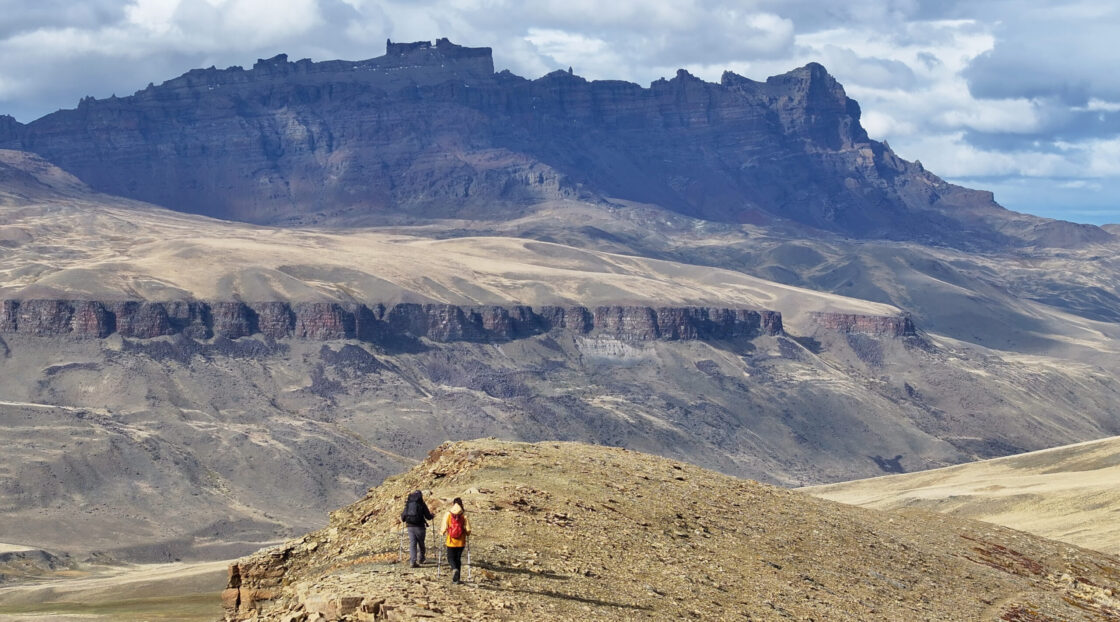 Screenshot Two hikers with trekking poles cross a rocky ridge beneath a vast, layered mountain range and cloudy sky.