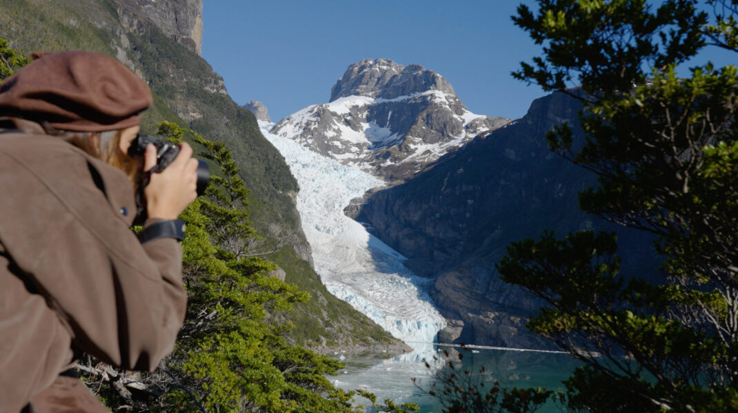 Hiker photographs a snow-covered mountain glacier descending into a turquoise lake in a rugged valley.
