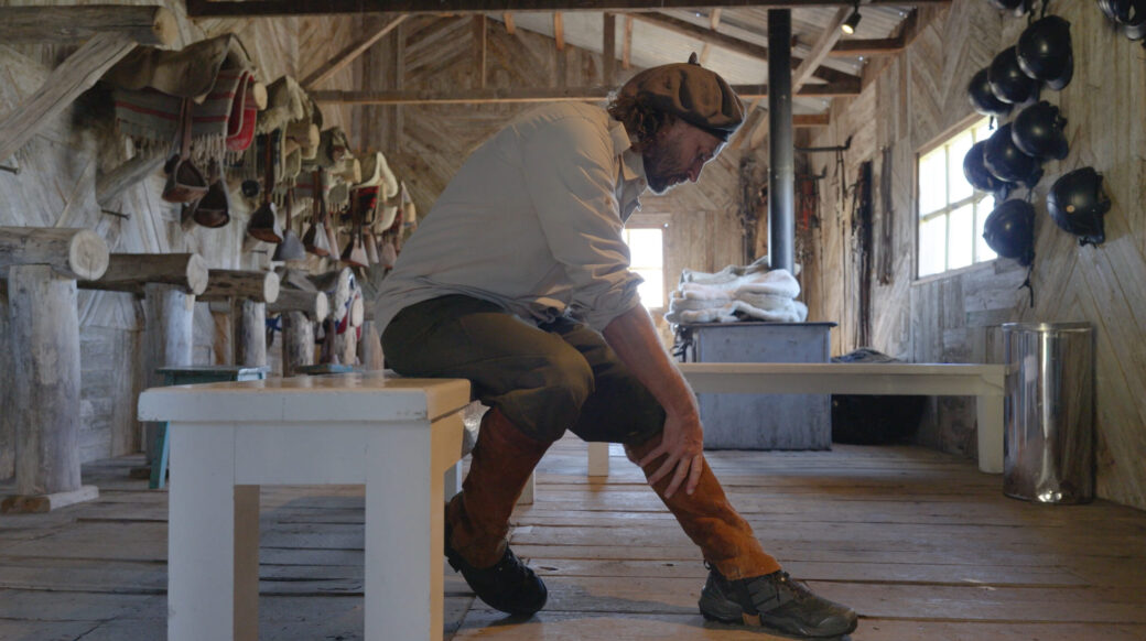 Bearded man in a rustic workshop bends to adjust his boot, surrounded by riding gear and helmets.