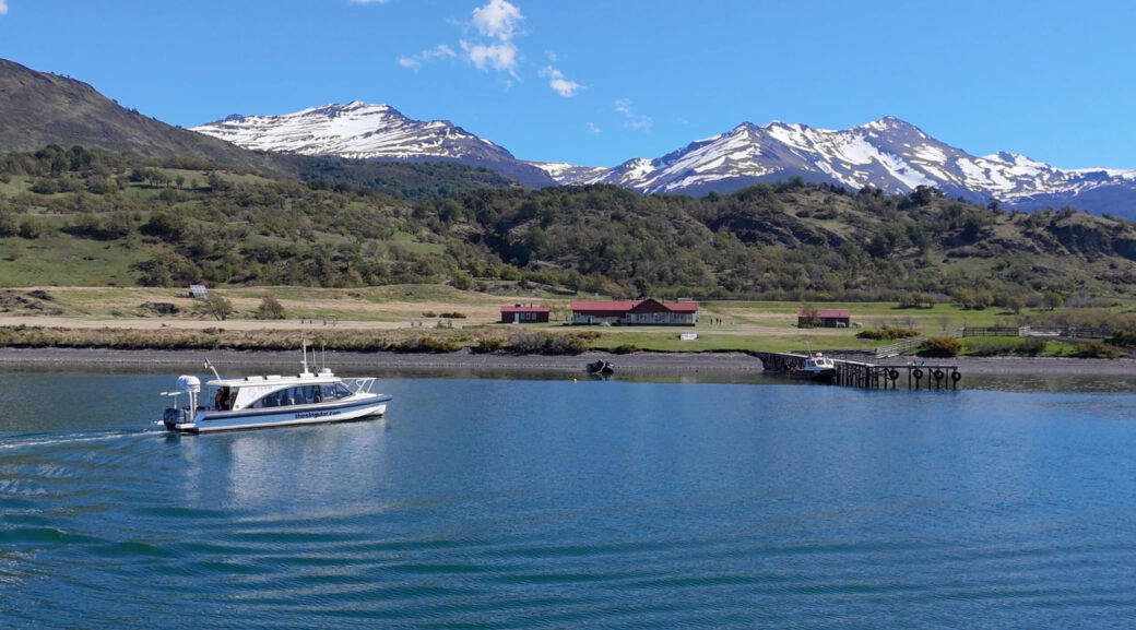 A white tour boat crosses a blue lake beneath snow-capped mountains, with a small lakeside settlement and pier on shore.