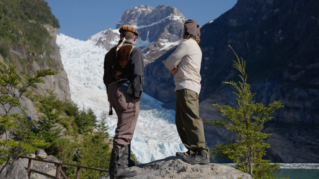 Two hikers admire a massive glacier beneath snow-covered mountains in a rugged alpine valley.