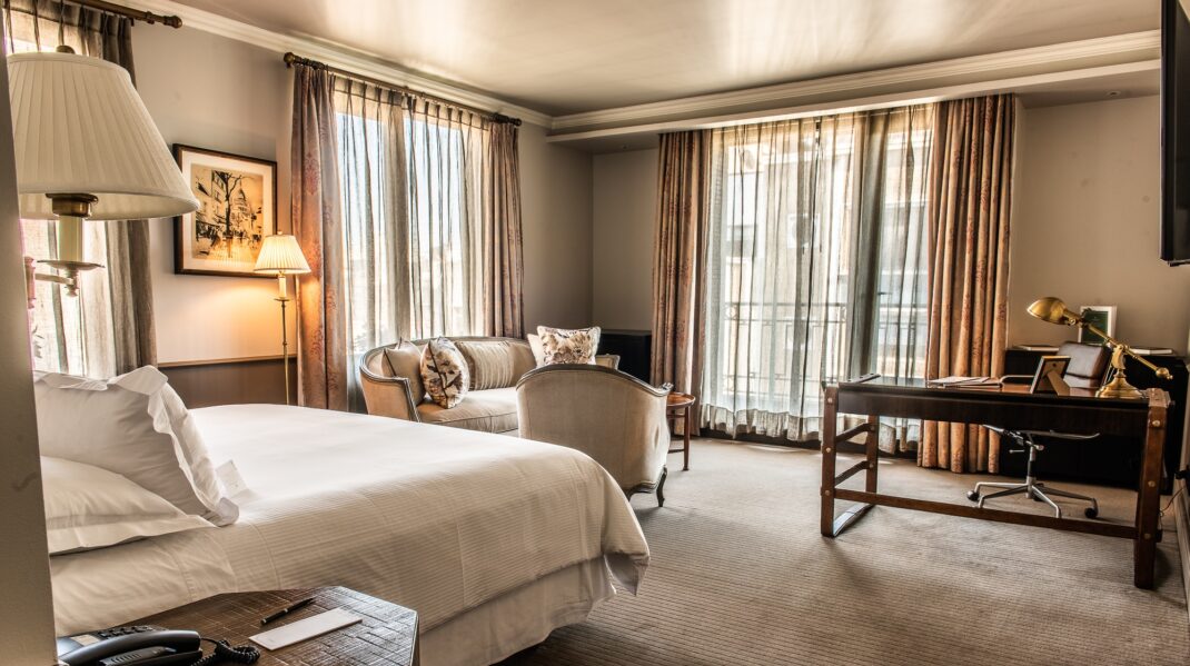 secrooms1 Elegant hotel suite with a king bed, sitting area, writing desk, and floor-to-ceiling curtained windows.